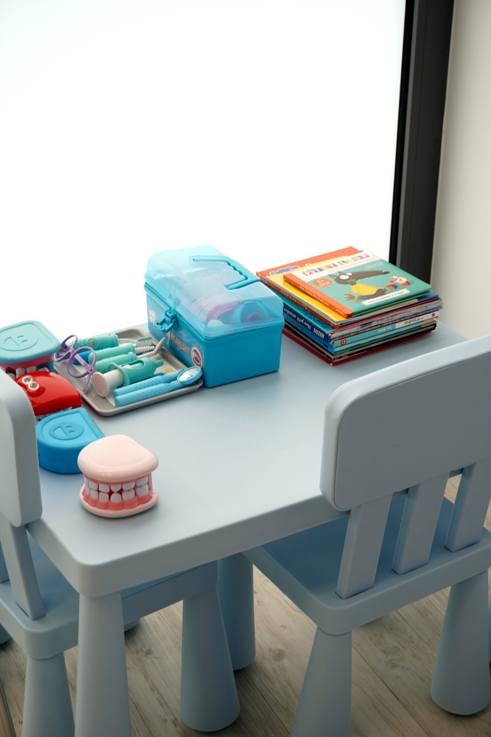 Espace enfants d’un cabinet dentaire avec petite table bleue, jeux de dentiste, mâchoire pédagogique et pile de livres illustrés.