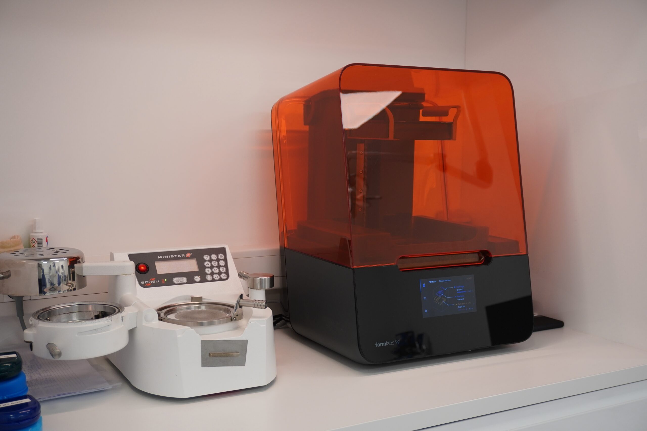Imprimante 3D dentaire avec capot orange translucide posée sur un plan de travail, à côté d’un appareil de thermoformage pour gouttières.