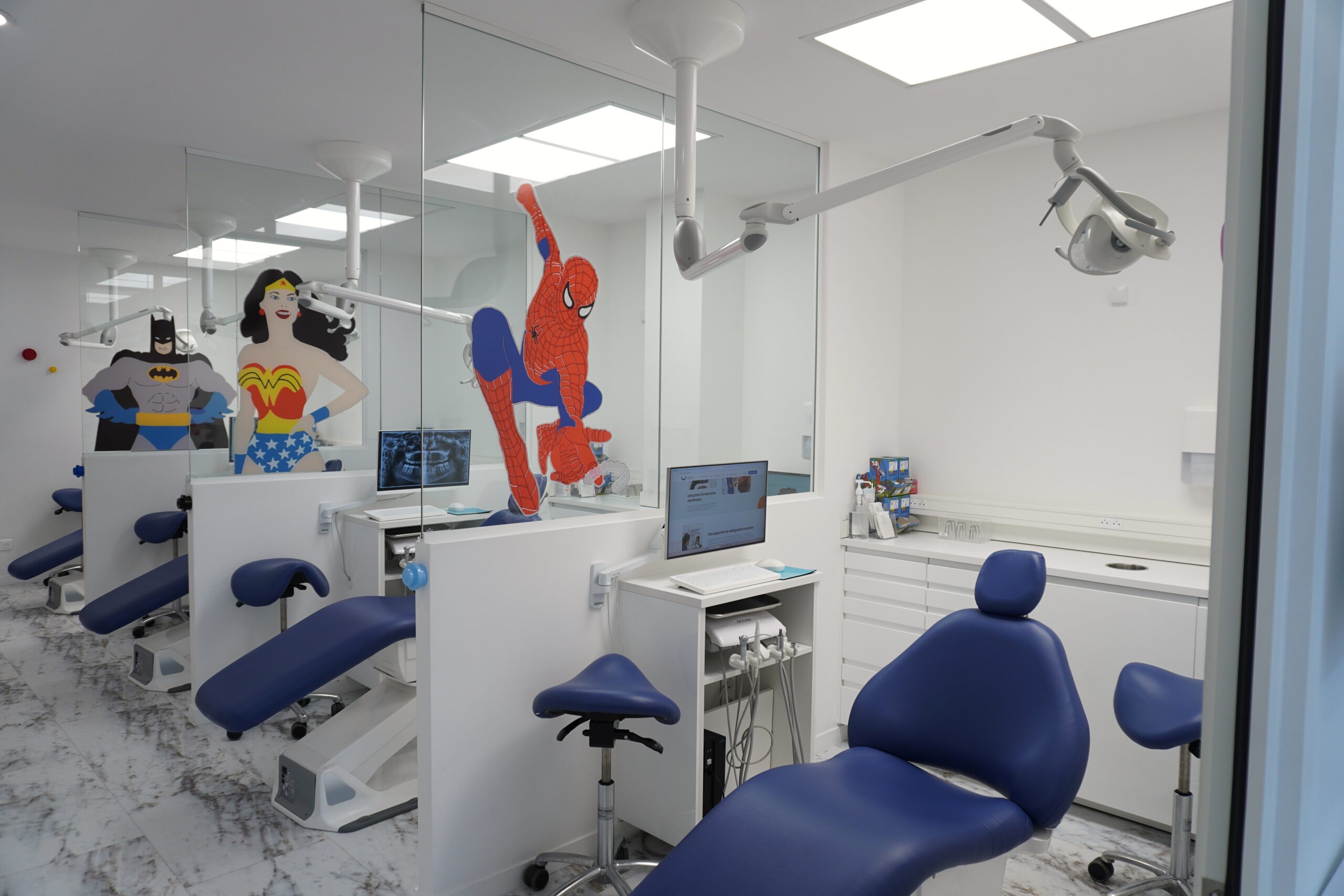 Salle de soins d’un cabinet dentaire avec fauteuils bleus, équipements médicaux et décorations murales de super-héros visibles dans le miroir.