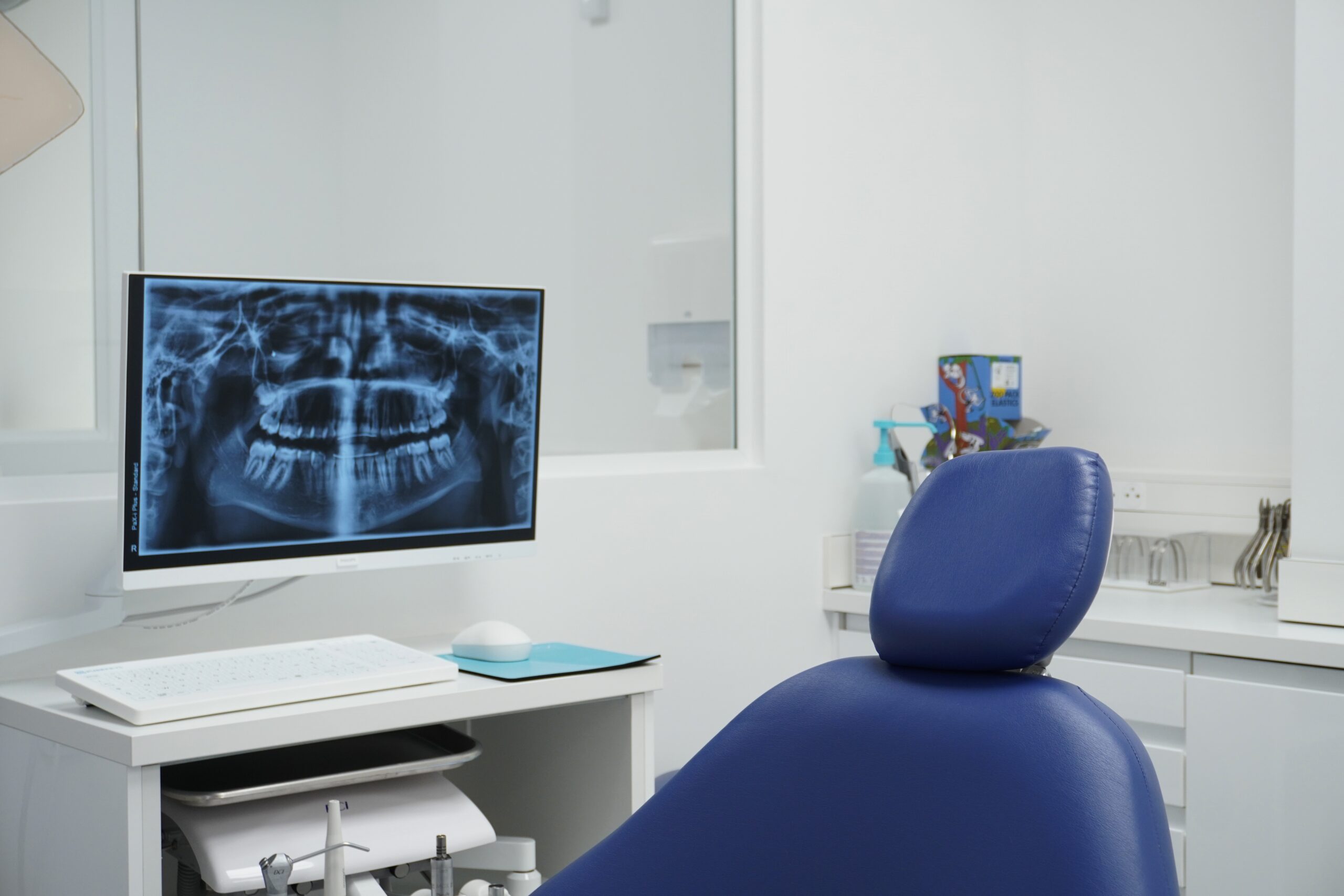 Cabinet dentaire moderne avec un fauteuil bleu au premier plan et un écran affichant une radiographie panoramique des dents.